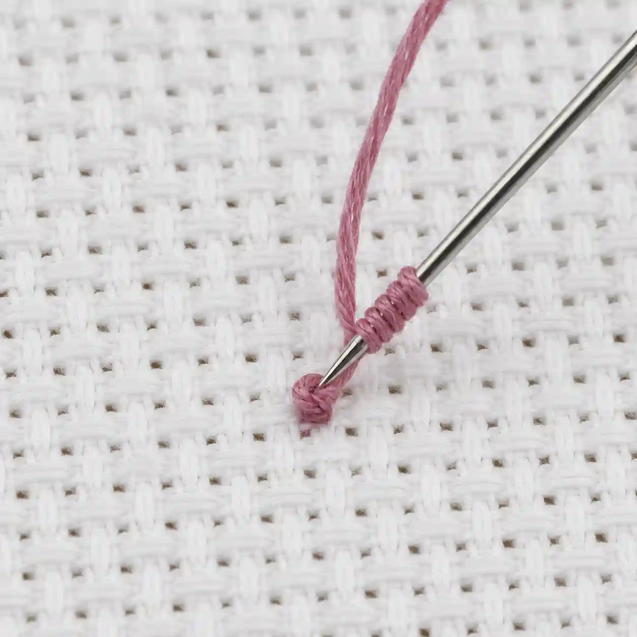 A needle wrapped with thread mid-stitch, forming a French knot on Aida fabric